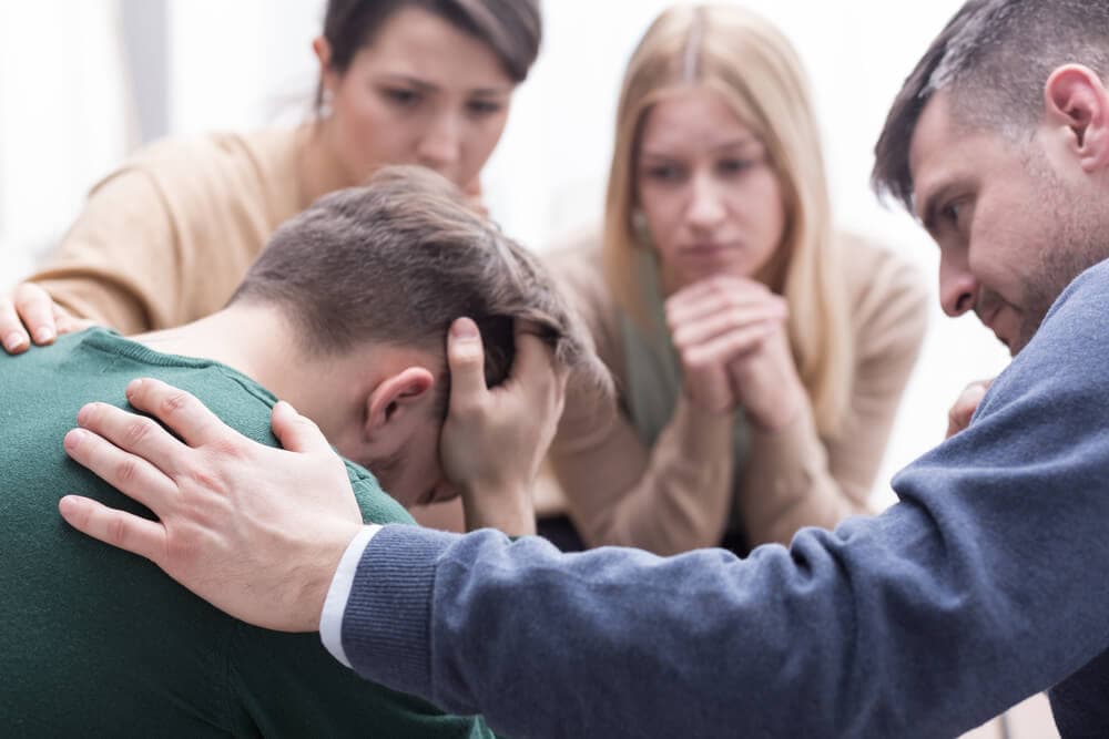 man with head in hands being consoled by a circle of concerned people